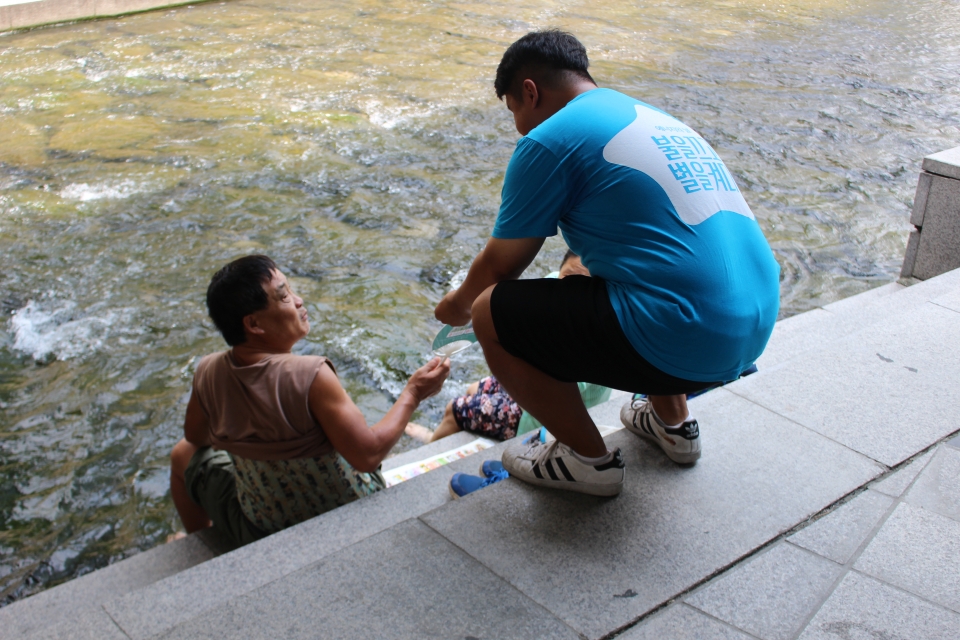 대학생 자원활동가가 청계천 다리 밑에서 휴식을 취하는 시민에게 홍보용 부채를 전달하고 있다.